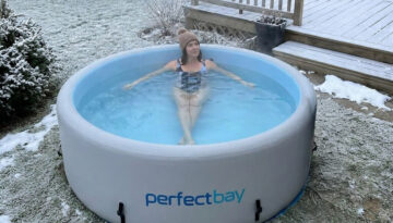Eine Frau in einem Badeanzug und einer Mütze sitzt im Cold Plunge Pool, der im Garten aufgestellt ist